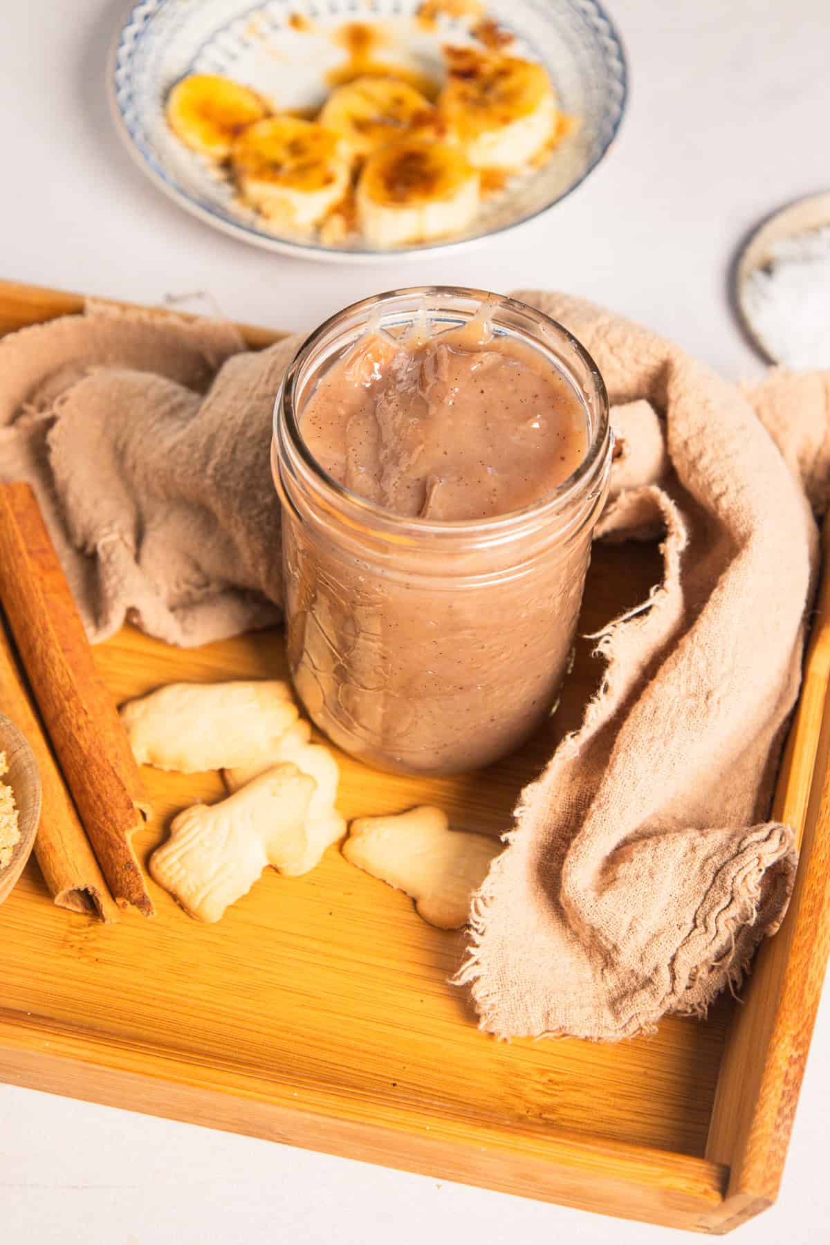 A glass jar of beige pudding with banana coffee syrup, animal cookies, and a cinnamon stick on a wooden tray.
