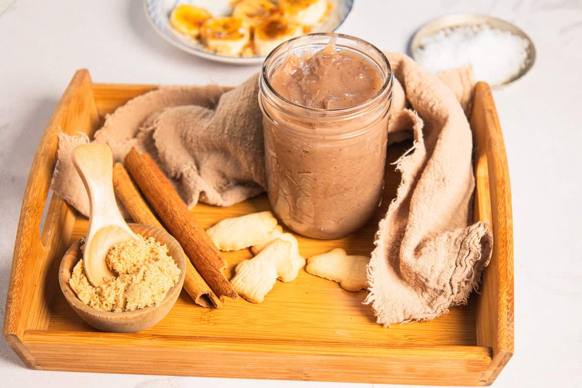 A jar of banana coffee syrup on a wooden tray with cinnamon sticks, brown sugar, and small cookies.