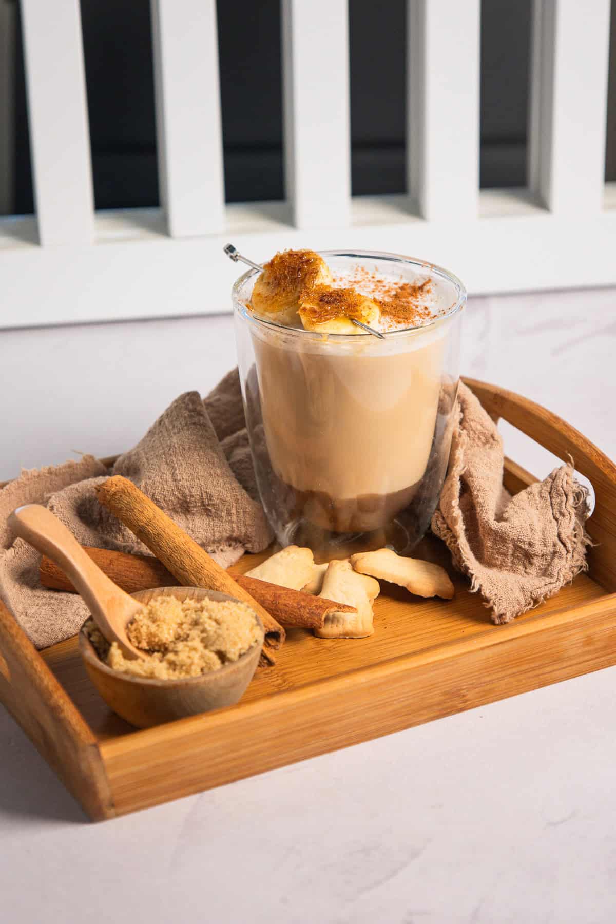 Iced Banana Bread Latte topped with banana slices, served on a wooden tray with cinnamon sticks, sugar, and cookies.