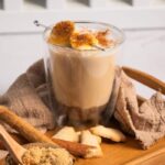 An Iced Banana Bread Latte topped with brûléed banana slices on a wooden tray with brown sugar and cinnamon.