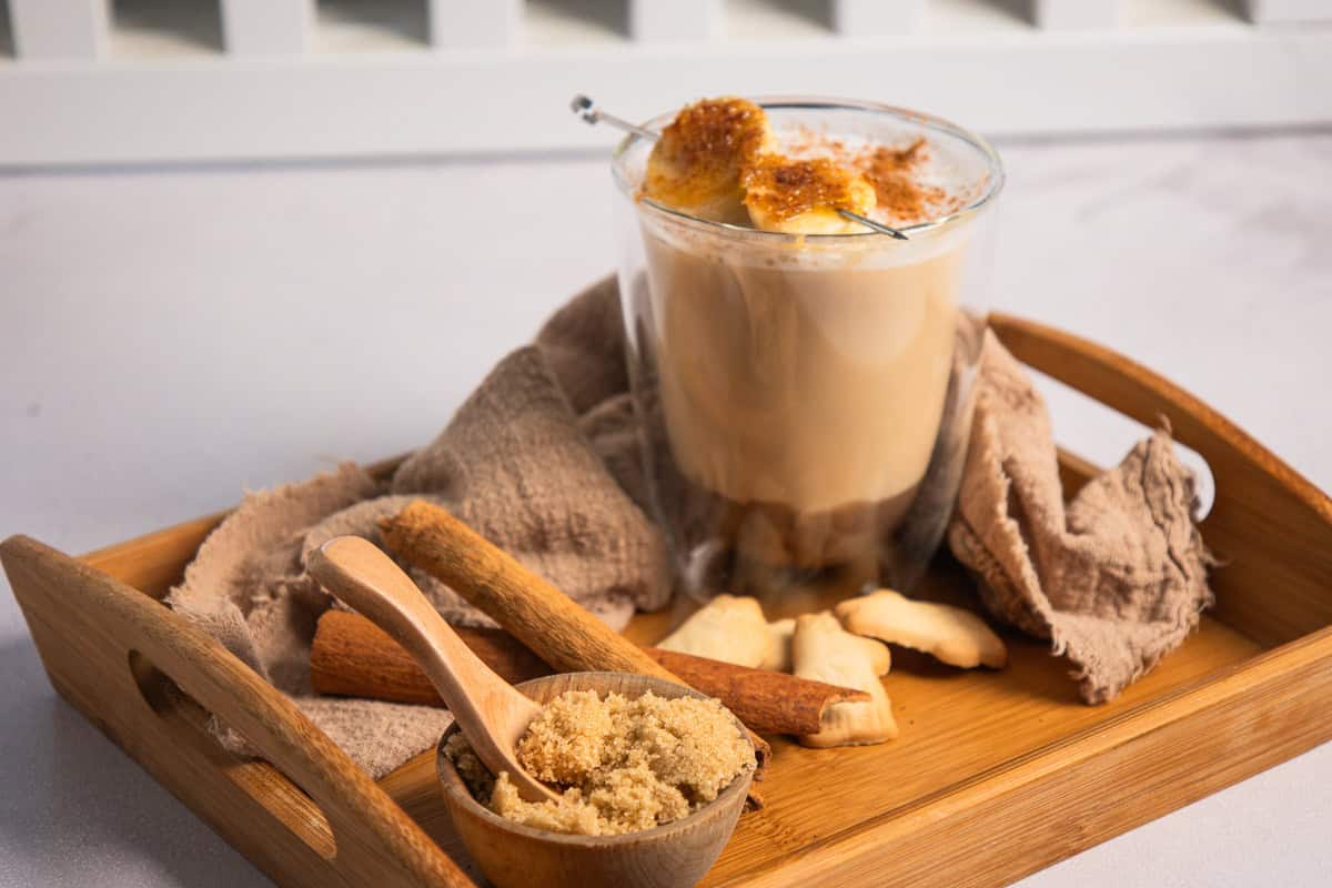 Glass of Iced Banana Bread Latte topped with br&ucirc;l&eacute;ed banana slices, served on a wooden tray with cinnamon sticks.