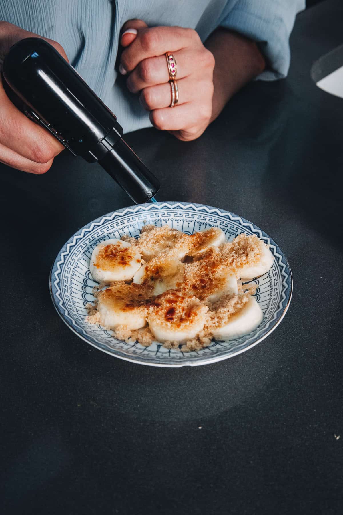 A hand uses a kitchen torch to caramelize brown sugar on sliced bananas for an iced banana bread latte.