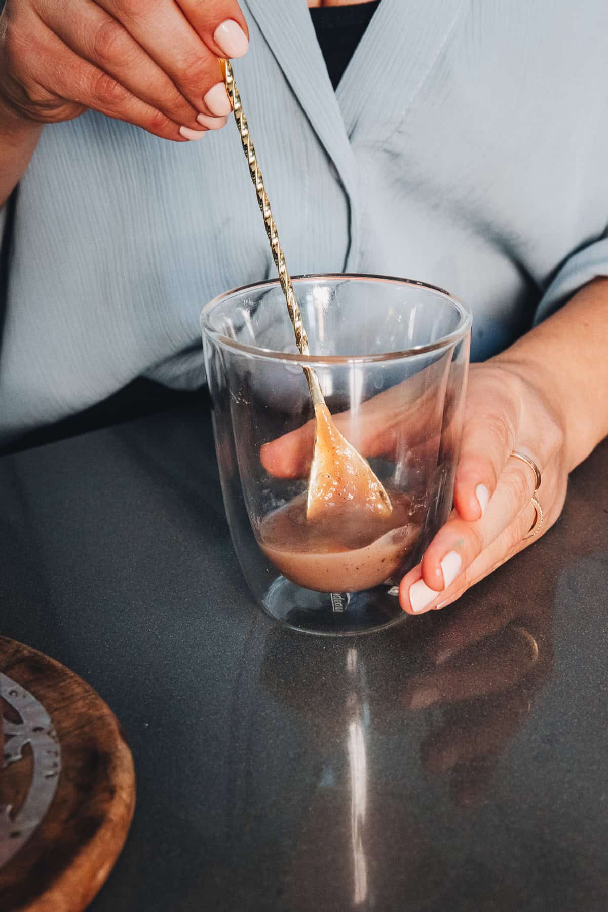 A person uses a long gold spoon to stir an Iced Banana Bread Latte in a double-walled glass.