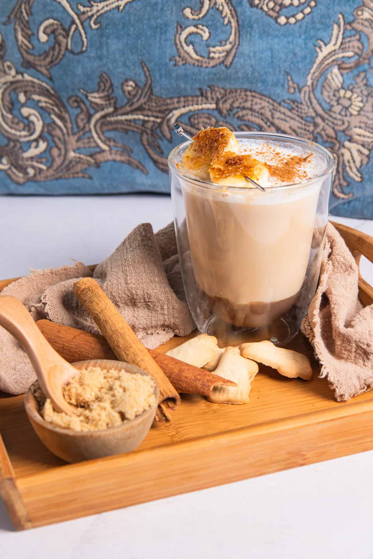 An Iced Banana Bread Latte topped with torched banana slices on a wooden tray with cinnamon, brown sugar, and crackers.