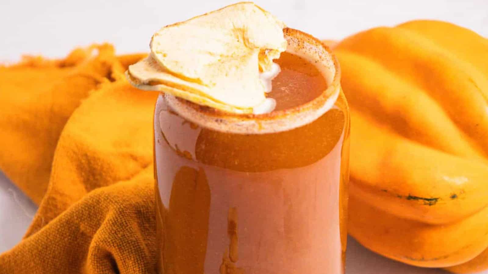 A glass of alcohol-free orange smoothie topped with dried apple slices, beside a squash and an orange cloth.