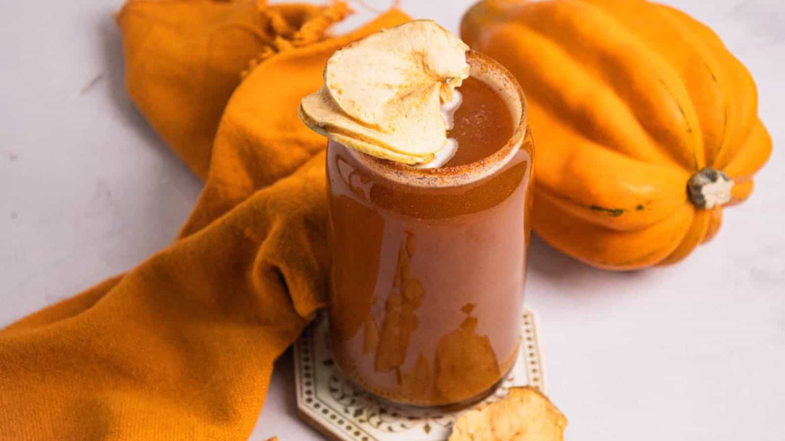 A glass of brown juice with apple chips, next to an orange cloth and a pumpkin on a light surface.