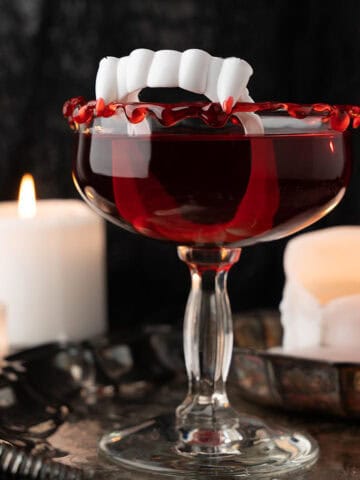 A red cocktail in a coupe glass rimmed with fake vampire fangs, surrounded by lit candles.